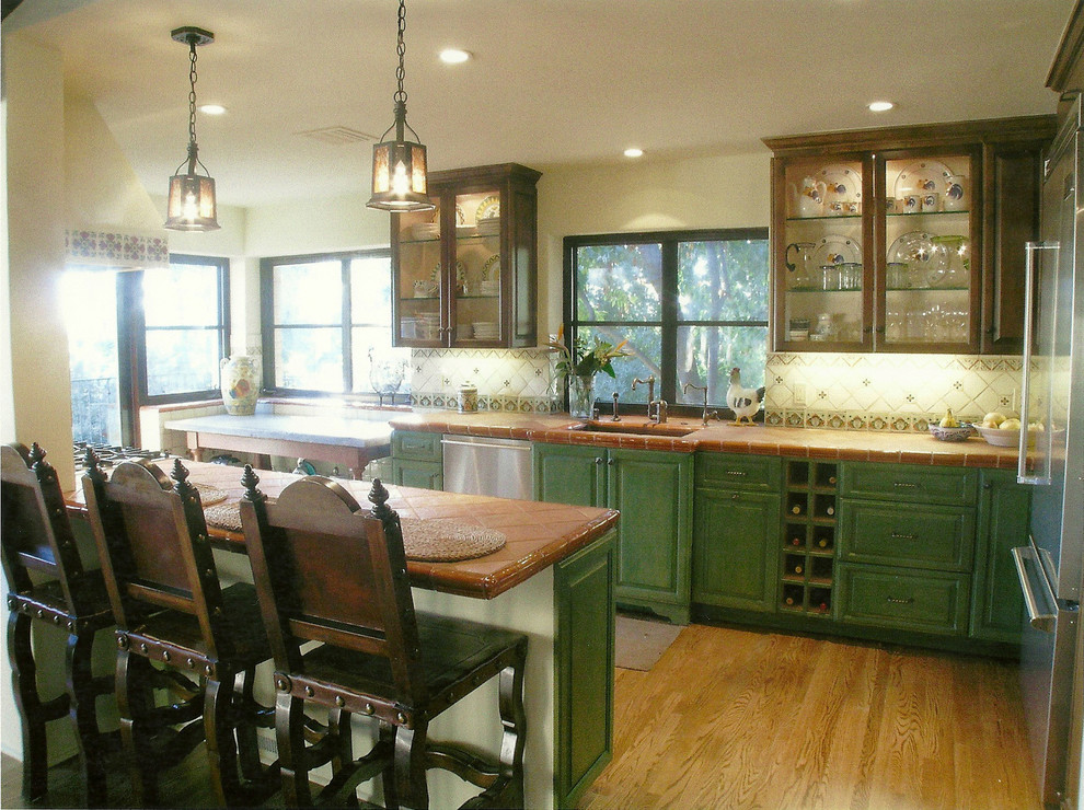 Example of a large tuscan medium tone wood floor kitchen design in Los Angeles with a peninsula, glass-front cabinets, dark wood cabinets, tile countertops, white backsplash, ceramic backsplash, stainless steel appliances and a drop-in sink
