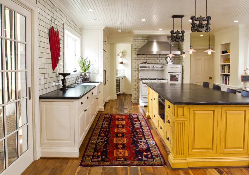 Lantlig inredning av ett mellanstort l-kök, med en rustik diskho, luckor med upphöjd panel, gula skåp, vitt stänkskydd, stänkskydd i tunnelbanekakel, vita vitvaror, mellanmörkt trägolv, en köksö och brunt golv