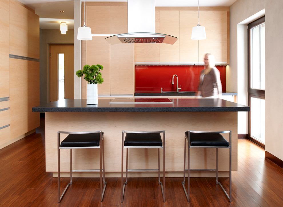 Réalisation d'une cuisine encastrable design en bois clair avec un placard à porte plane, une crédence rouge, une crédence en feuille de verre, parquet foncé et îlot.