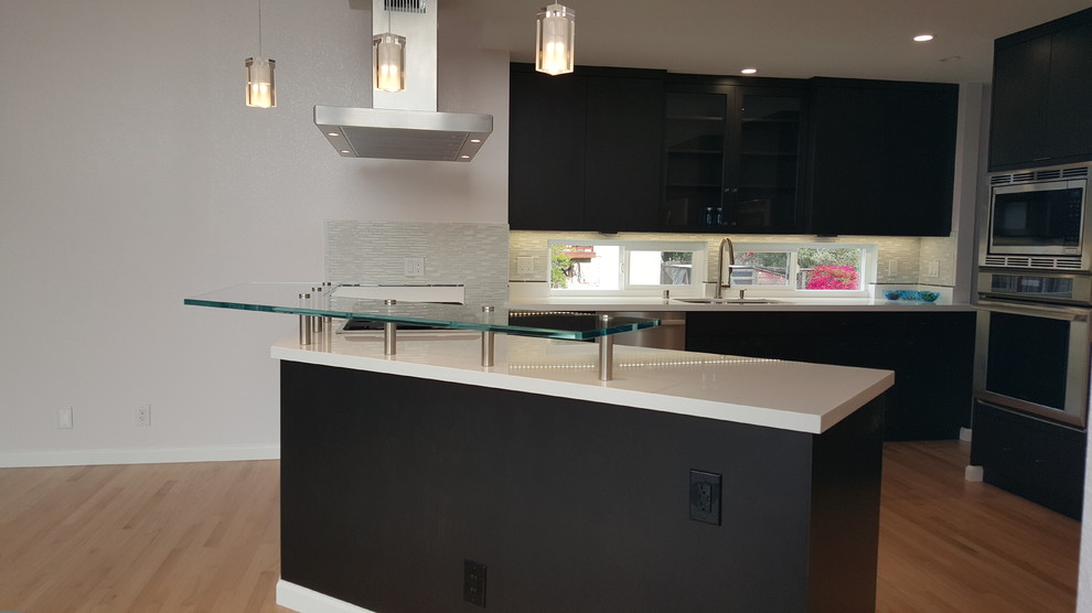 Medium sized modern u-shaped kitchen/diner in San Francisco with a submerged sink, flat-panel cabinets, black cabinets, engineered stone countertops, grey splashback, glass tiled splashback, stainless steel appliances, light hardwood flooring, a breakfast bar and brown floors.