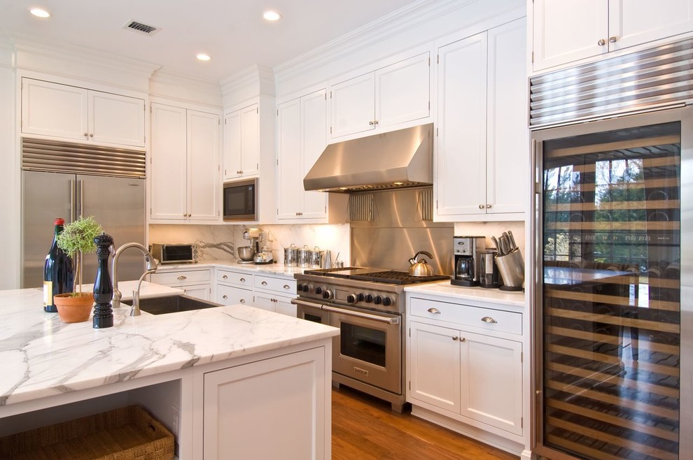 Large transitional l-shaped medium tone wood floor and brown floor eat-in kitchen photo in Other with a farmhouse sink, recessed-panel cabinets, white cabinets, marble countertops, white backsplash, marble backsplash, stainless steel appliances and an island