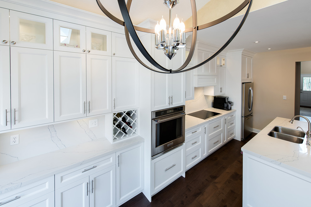 Large traditional single-wall kitchen/diner in Toronto with a double-bowl sink, shaker cabinets, white cabinets, engineered stone countertops, white splashback, stone slab splashback, stainless steel appliances, medium hardwood flooring, an island and brown floors.