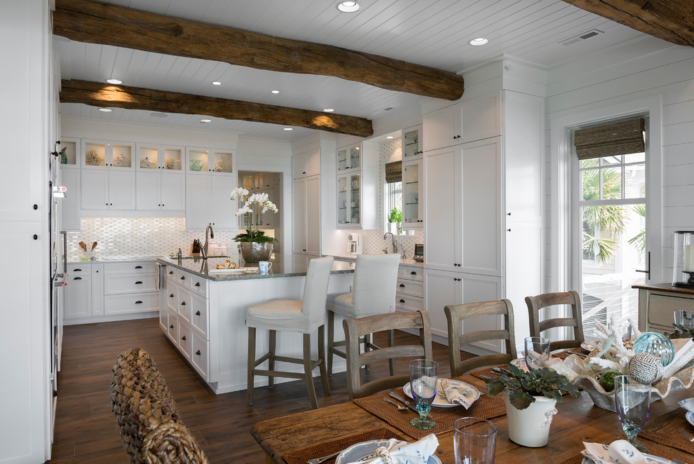 Aménagement d'une cuisine américaine bord de mer avec un placard à porte shaker, des portes de placard blanches, une crédence blanche, parquet foncé et îlot.