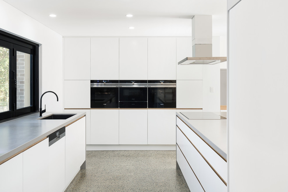Modern inredning av ett stort vit vitt kök, med en undermonterad diskho, vita skåp, bänkskiva i koppar, vitt stänkskydd, fönster som stänkskydd, svarta vitvaror, betonggolv, en köksö och grått golv