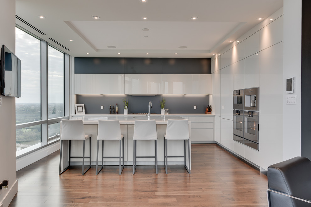 Design ideas for an expansive contemporary l-shaped open plan kitchen in Calgary with a submerged sink, flat-panel cabinets, white cabinets, engineered stone countertops, grey splashback, stainless steel appliances, medium hardwood flooring, an island, stone slab splashback and brown floors.