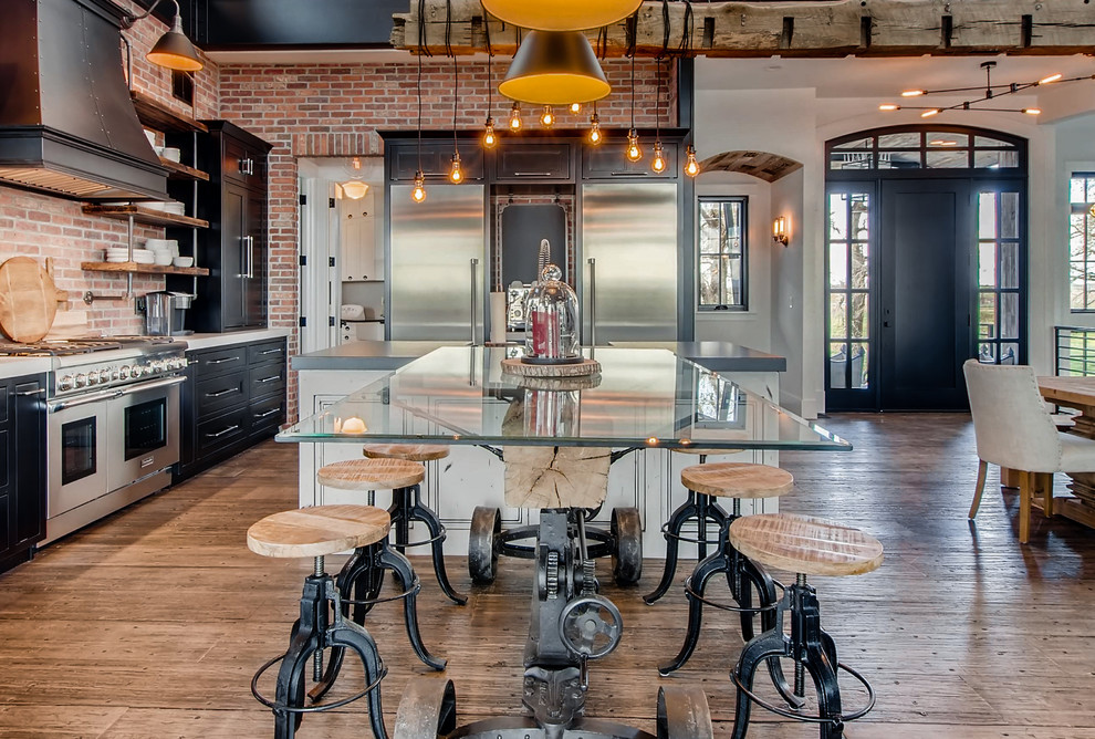 Cette image montre une cuisine américaine urbaine en L avec un placard à porte shaker, des portes de placard noires, une crédence en brique, un électroménager en acier inoxydable, un sol en bois brun et un sol marron.