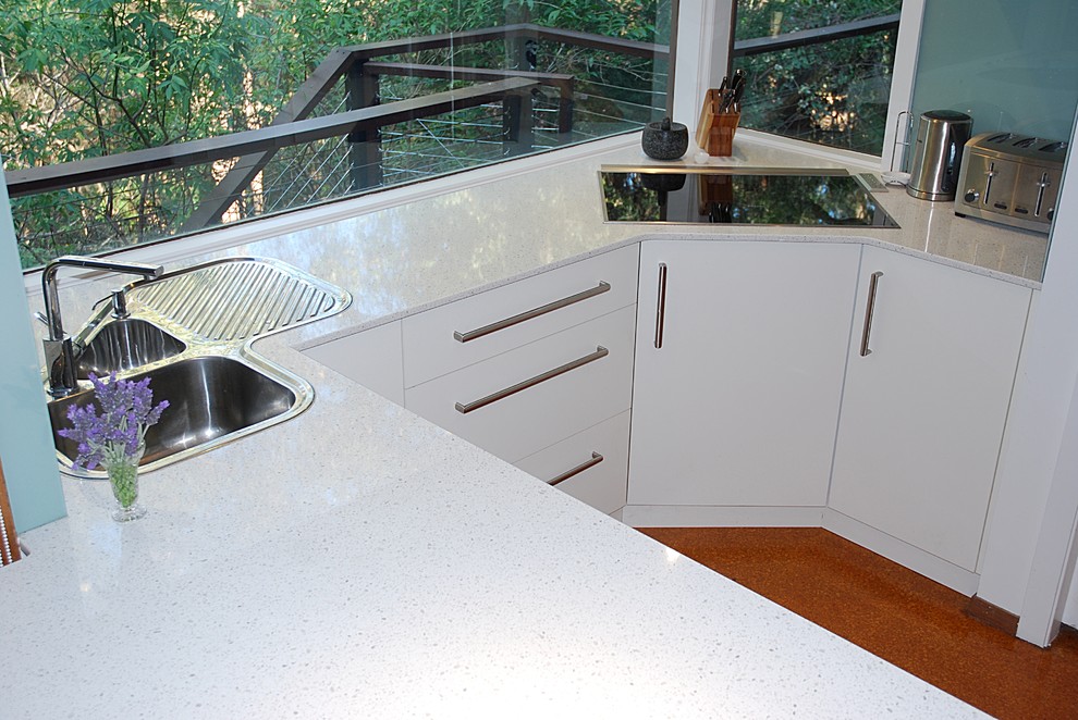 Photo of a medium sized modern u-shaped kitchen pantry in Sydney with a built-in sink, flat-panel cabinets, white cabinets, engineered stone countertops, green splashback, glass sheet splashback, stainless steel appliances, cork flooring and a breakfast bar.