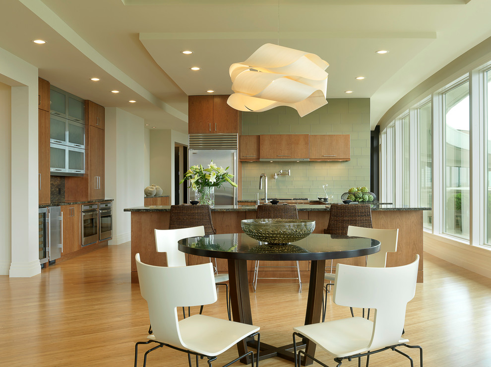 На фото: кухня в стиле модернизм с обеденным столом, плоскими фасадами, фасадами цвета дерева среднего тона, зеленым фартуком и техникой из нержавеющей стали