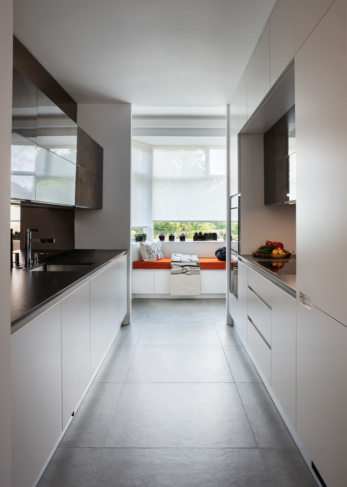 Inspiration for a contemporary galley kitchen in London with a submerged sink, flat-panel cabinets, white cabinets, mirror splashback, integrated appliances, grey floors and black worktops.