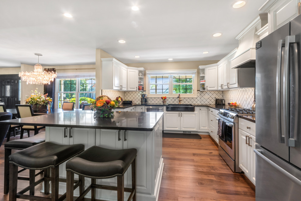 This is an example of a large traditional u-shaped kitchen/diner in Milwaukee with dark hardwood flooring, brown floors, a belfast sink, raised-panel cabinets, white cabinets, engineered stone countertops, white splashback, ceramic splashback, stainless steel appliances, an island and black worktops.