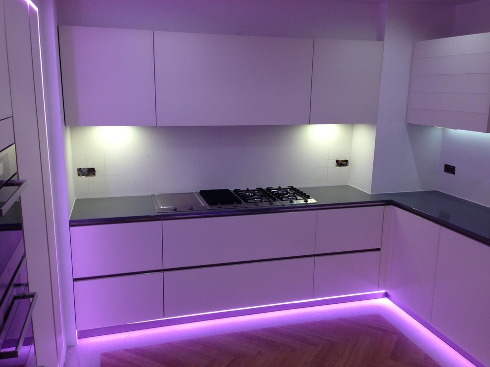 This is an example of a large contemporary u-shaped kitchen/diner in West Midlands with a submerged sink, glass-front cabinets, beige cabinets, glass worktops, white splashback, glass sheet splashback, stainless steel appliances, light hardwood flooring and no island.