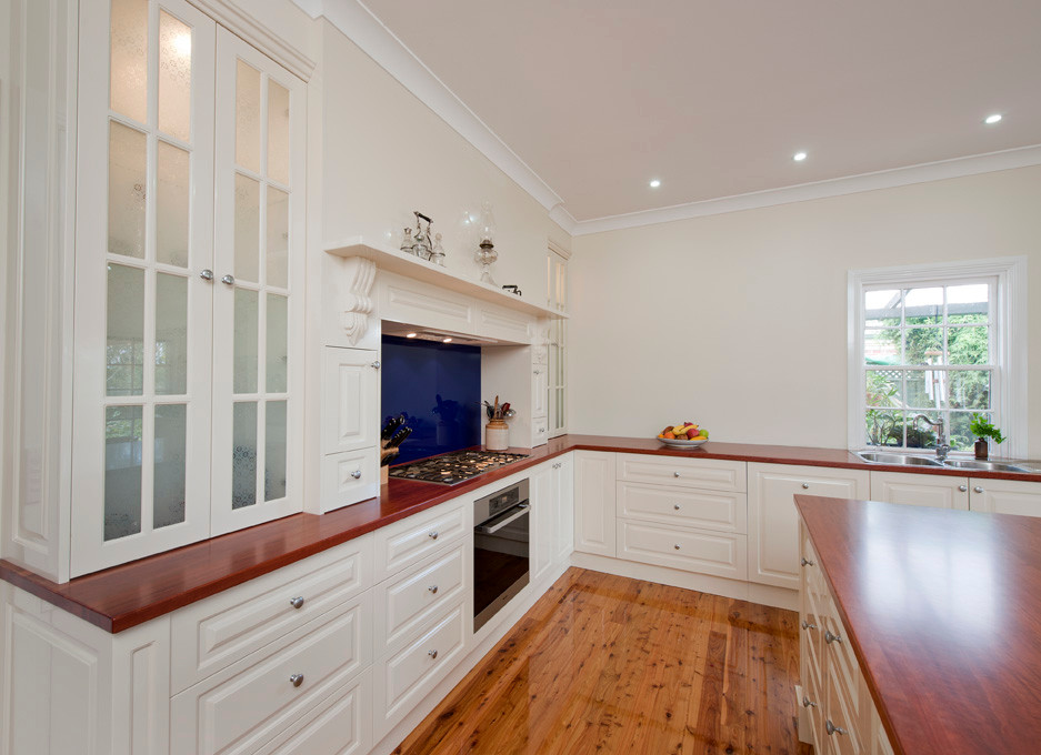 Inspiration for a large classic single-wall kitchen in Melbourne with a double-bowl sink, white cabinets, wood worktops, blue splashback, glass sheet splashback, stainless steel appliances, medium hardwood flooring and an island.