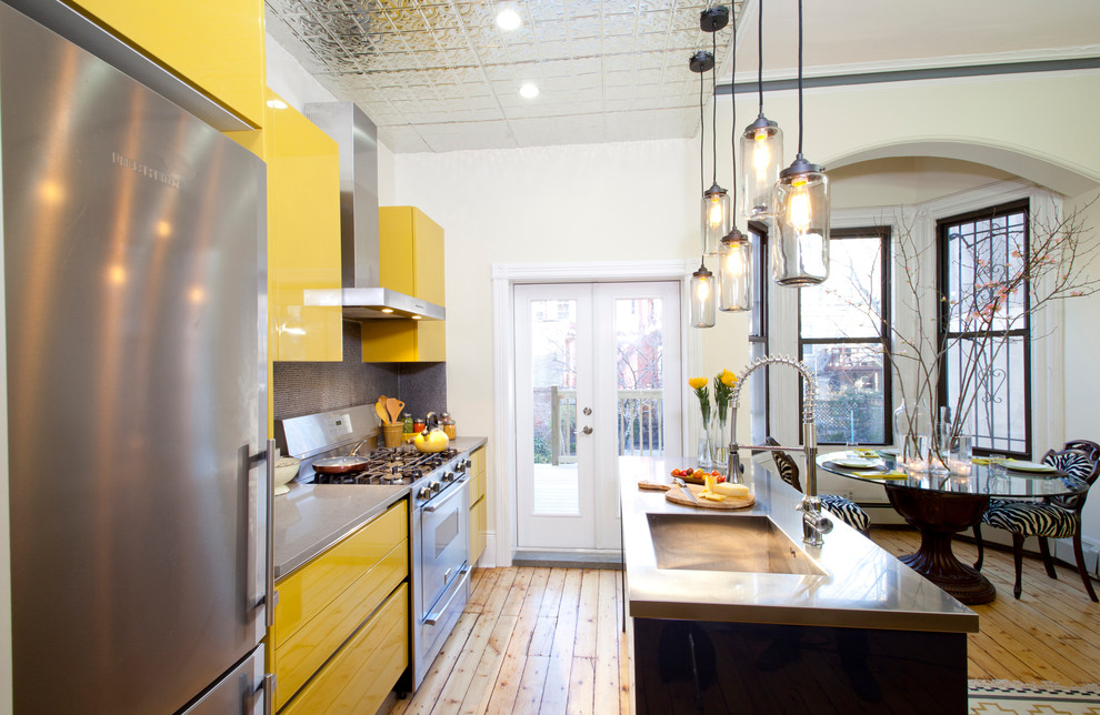 Cette image montre une cuisine américaine parallèle design avec un évier intégré, un placard à porte plane, des portes de placard jaunes, un plan de travail en inox et un électroménager en acier inoxydable.