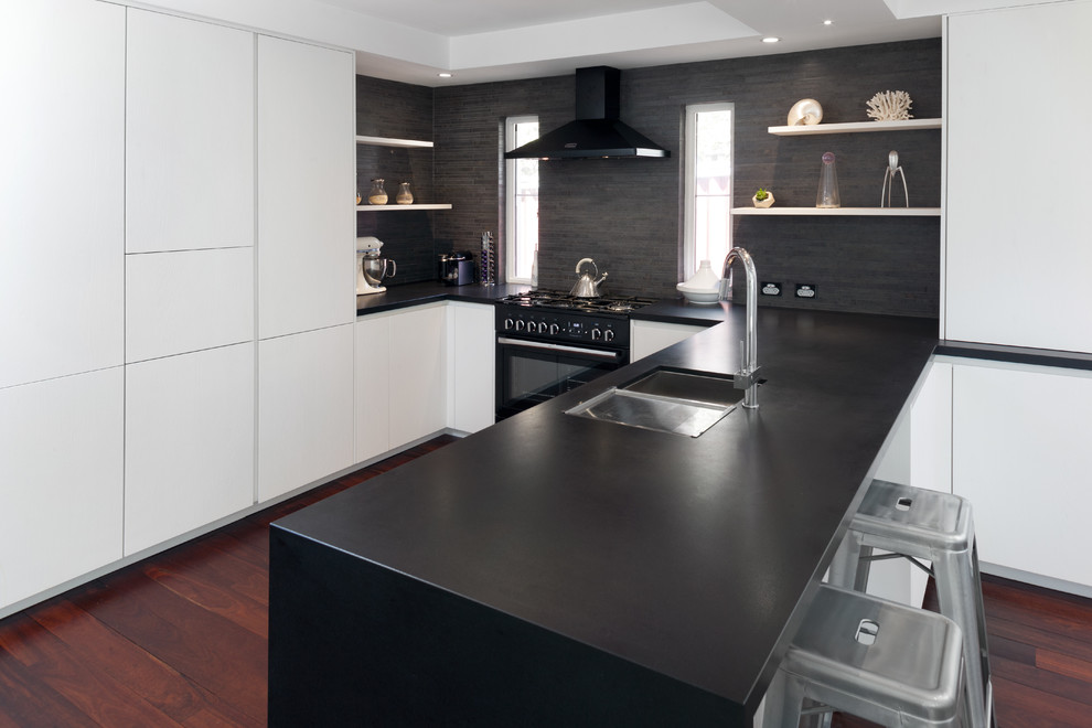 Example of a large minimalist u-shaped medium tone wood floor eat-in kitchen design in Perth with white cabinets, granite countertops, black backsplash, ceramic backsplash, black appliances and an island