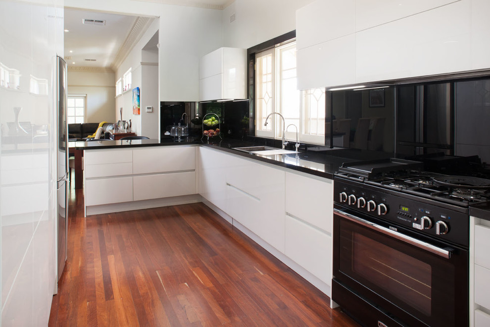 Mittelgroße Moderne Wohnküche in L-Form mit Doppelwaschbecken, weißen Schränken, Granit-Arbeitsplatte, Küchenrückwand in Schwarz, Rückwand aus Glasfliesen, schwarzen Elektrogeräten, braunem Holzboden und Kücheninsel in Perth