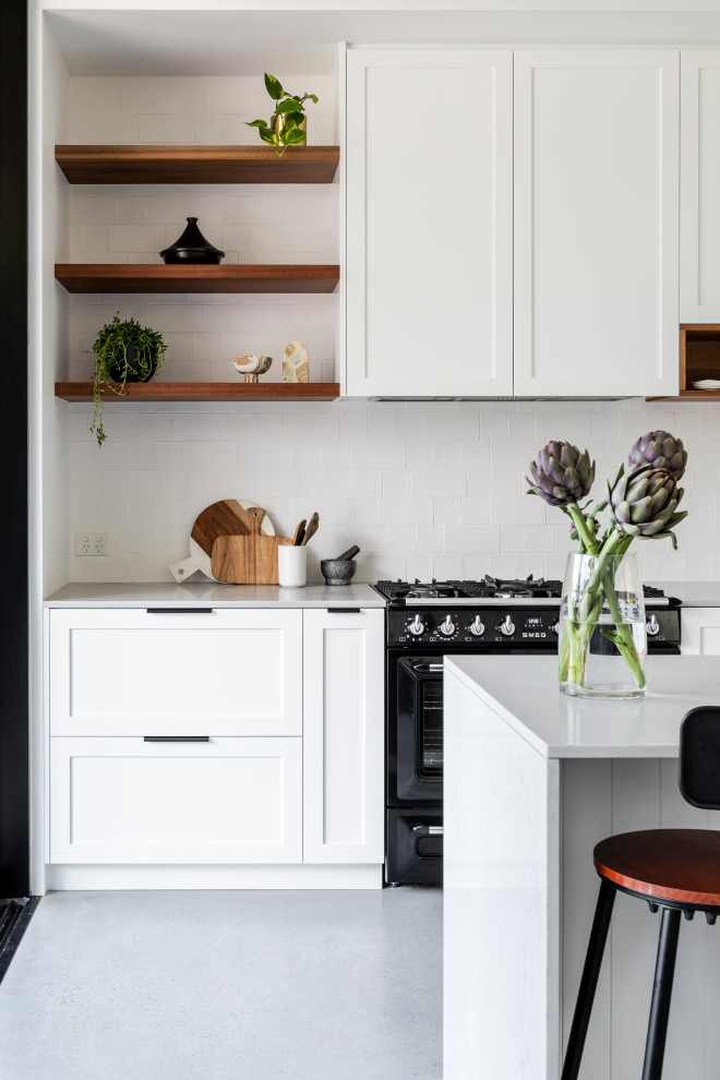 Bild på ett mellanstort vintage grå linjärt grått kök med öppen planlösning, med en undermonterad diskho, skåp i shakerstil, vita skåp, bänkskiva i kvarts, vitt stänkskydd, stänkskydd i keramik, svarta vitvaror, betonggolv, en köksö och grått golv