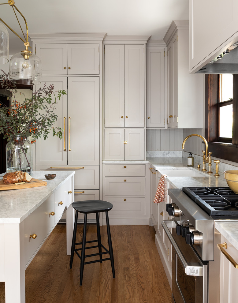 Inredning av ett klassiskt grå grått l-kök, med en rustik diskho, skåp i shakerstil, grå skåp, brunt stänkskydd, rostfria vitvaror, mellanmörkt trägolv, en köksö och brunt golv