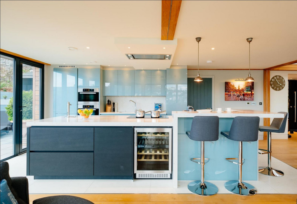 Идея дизайна: кухня-гостиная в современном стиле с плоскими фасадами, синими фасадами, белым фартуком, техникой из нержавеющей стали, островом и белой столешницей