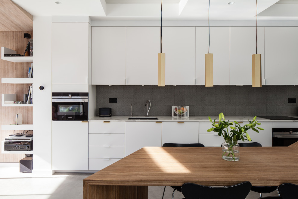 Modern inredning av ett vit linjärt vitt kök och matrum, med en undermonterad diskho, släta luckor, vita skåp, grått stänkskydd, stänkskydd i mosaik, rostfria vitvaror, betonggolv och grått golv