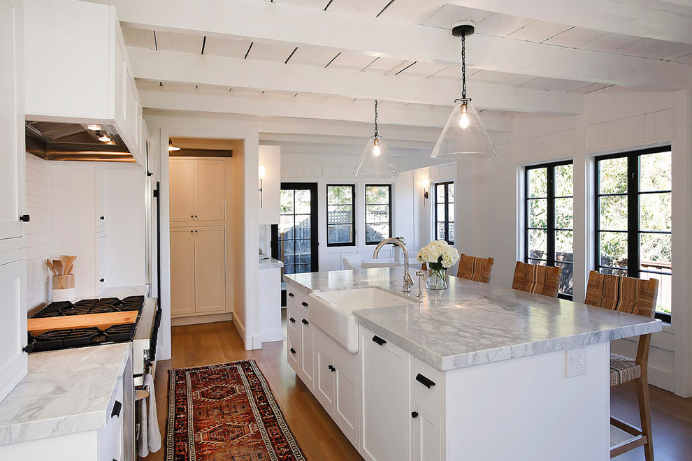 Photo of a medium sized nautical single-wall kitchen/diner in San Francisco with a belfast sink, shaker cabinets, white cabinets, marble worktops, white splashback, metro tiled splashback, integrated appliances, light hardwood flooring, an island, brown floors and white worktops.