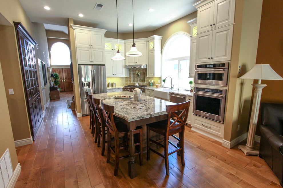 Exempel på ett mellanstort klassiskt kök, med en rustik diskho, luckor med upphöjd panel, vita skåp, granitbänkskiva, beige stänkskydd, stänkskydd i keramik, rostfria vitvaror, ljust trägolv och en köksö