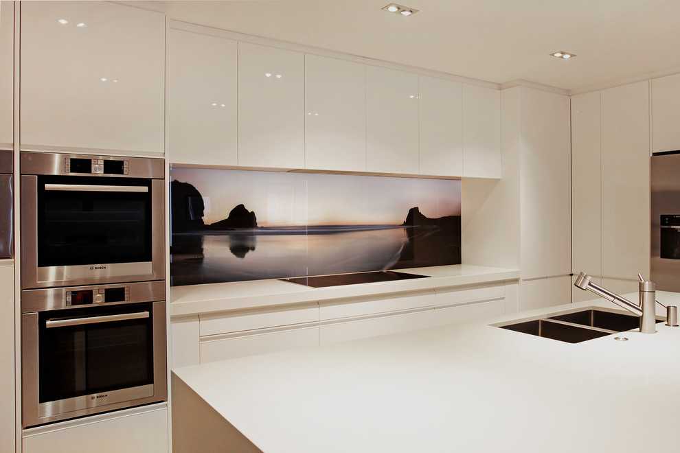Design ideas for a medium sized contemporary kitchen in Auckland with a double-bowl sink, recessed-panel cabinets, white cabinets, composite countertops, black splashback, glass sheet splashback, stainless steel appliances, light hardwood flooring and an island.