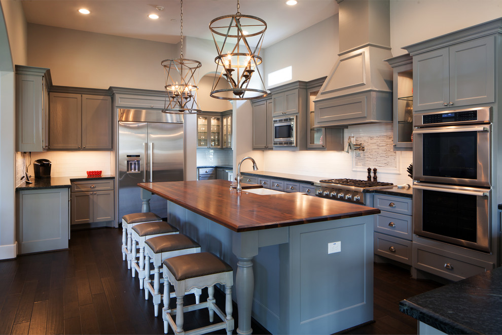 Klassische Küche in grau-weiß in U-Form mit Landhausspüle, Schrankfronten im Shaker-Stil, grauen Schränken, Arbeitsplatte aus Holz, Küchenrückwand in Weiß, Küchengeräten aus Edelstahl, dunklem Holzboden und Kücheninsel in Houston