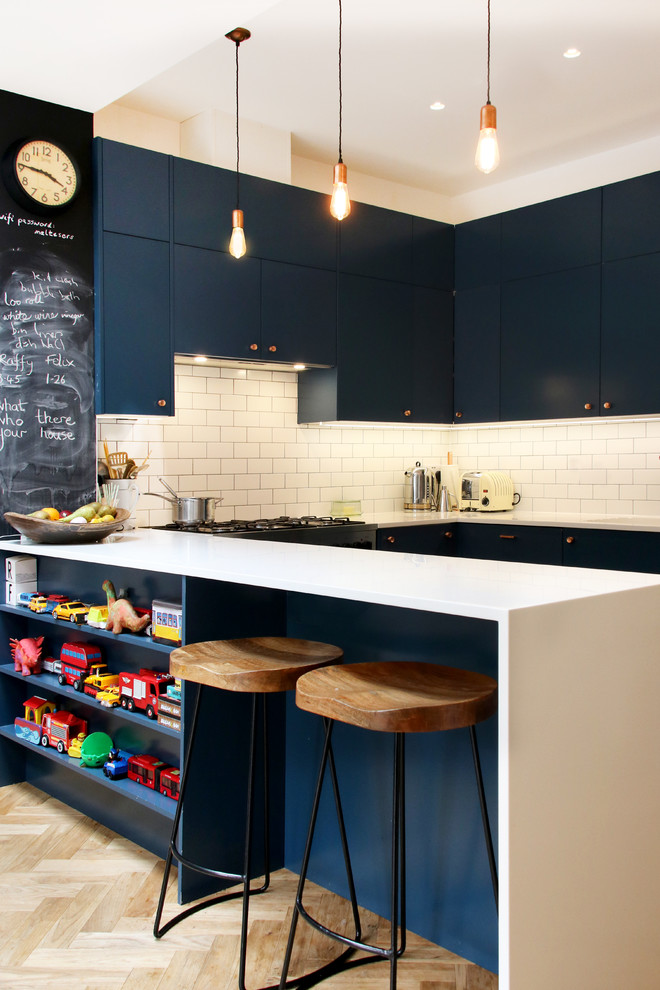 Trendy u-shaped light wood floor and beige floor kitchen photo in London with flat-panel cabinets, blue cabinets, white backsplash, subway tile backsplash, white countertops and a peninsula