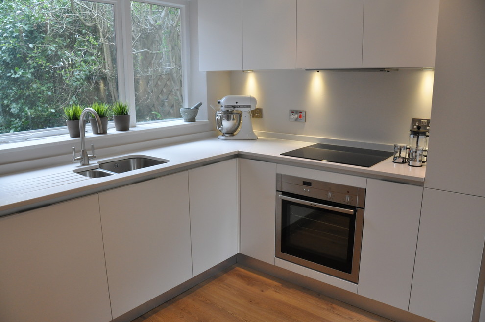 Design ideas for a small contemporary u-shaped kitchen/diner in Cheshire with a built-in sink, flat-panel cabinets, white cabinets, composite countertops, white splashback, glass sheet splashback, stainless steel appliances, medium hardwood flooring and a breakfast bar.