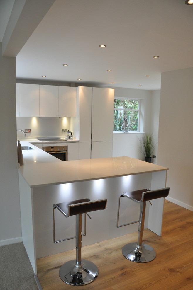 This is an example of a small contemporary u-shaped kitchen/diner in Cheshire with a built-in sink, flat-panel cabinets, white cabinets, composite countertops, white splashback, glass sheet splashback, stainless steel appliances, medium hardwood flooring and a breakfast bar.