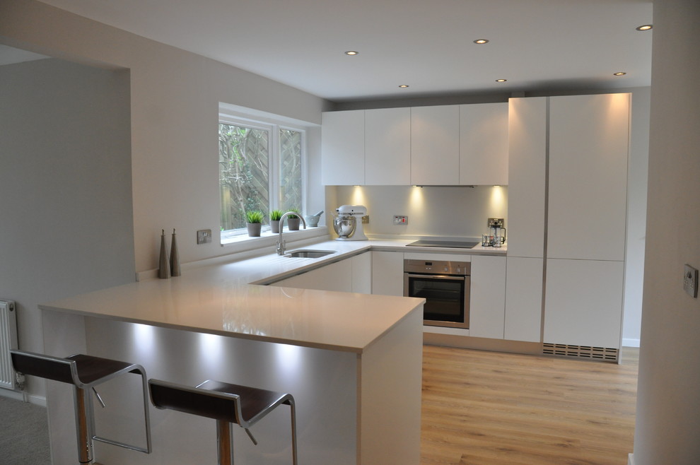 Inspiration for a small contemporary u-shaped kitchen/diner in Cheshire with a built-in sink, flat-panel cabinets, white cabinets, composite countertops, white splashback, glass sheet splashback, stainless steel appliances, medium hardwood flooring and a breakfast bar.