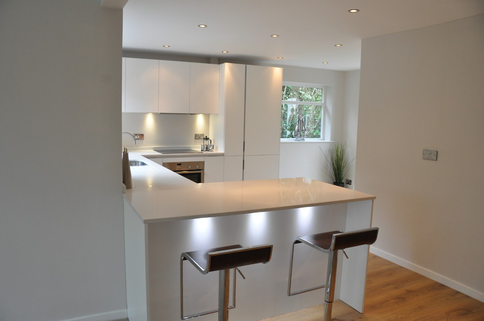 Example of a small trendy u-shaped medium tone wood floor eat-in kitchen design in Cheshire with a drop-in sink, flat-panel cabinets, white cabinets, solid surface countertops, white backsplash, glass sheet backsplash, stainless steel appliances and a peninsula