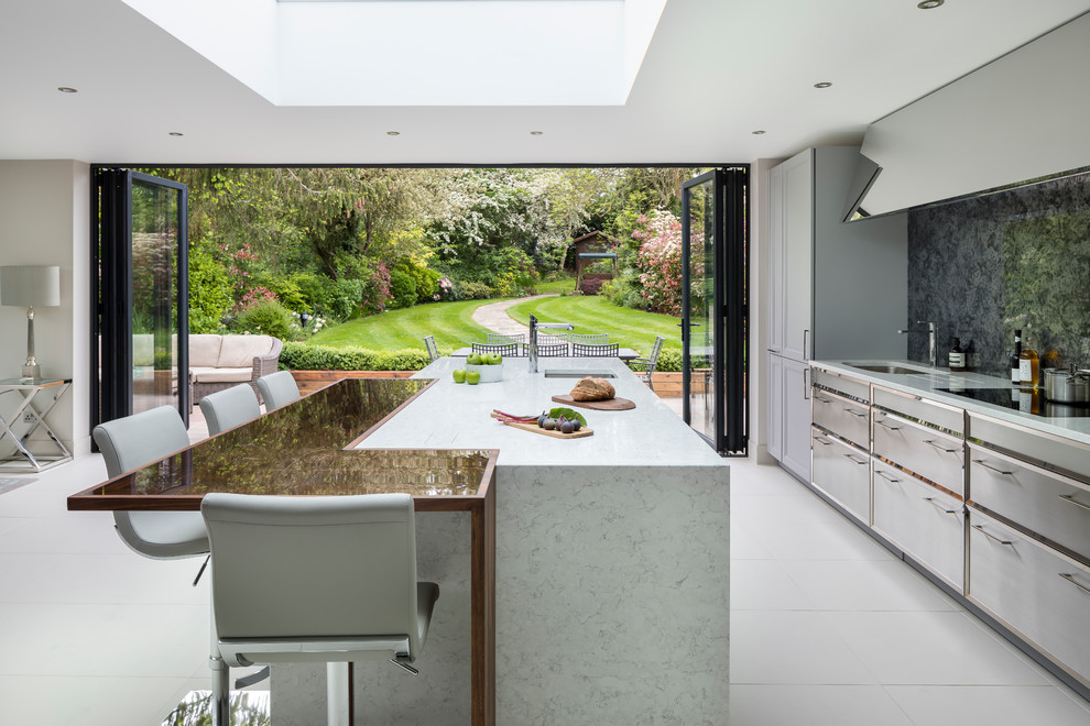 This is an example of a large traditional single-wall open plan kitchen in London with a built-in sink, recessed-panel cabinets, grey cabinets, composite countertops, metallic splashback, mirror splashback, black appliances, ceramic flooring, an island and white floors.