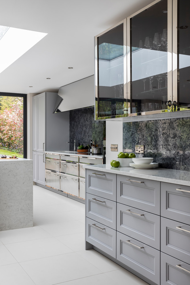 Photo of a large traditional single-wall open plan kitchen in London with a built-in sink, recessed-panel cabinets, grey cabinets, composite countertops, metallic splashback, mirror splashback, black appliances, ceramic flooring, an island and white floors.