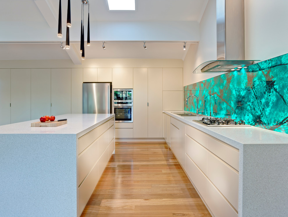 Inspiration for an expansive contemporary open plan kitchen in Other with a built-in sink, green splashback, glass tiled splashback, stainless steel appliances, light hardwood flooring and an island.