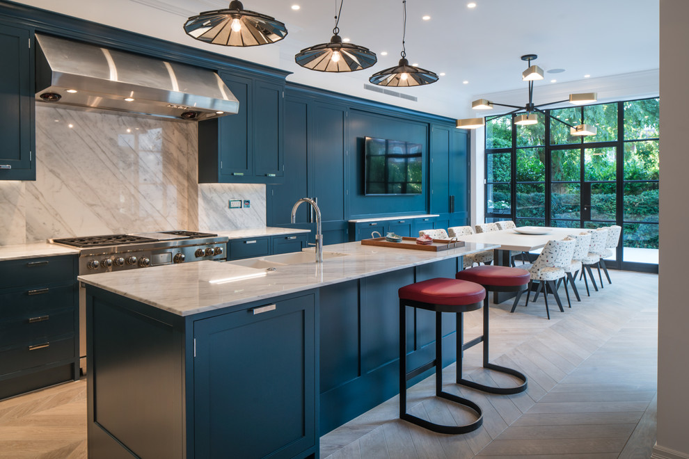 Photo of a medium sized traditional kitchen/diner in London with blue cabinets, marble worktops, marble splashback, light hardwood flooring, an island, shaker cabinets, white splashback and stainless steel appliances.