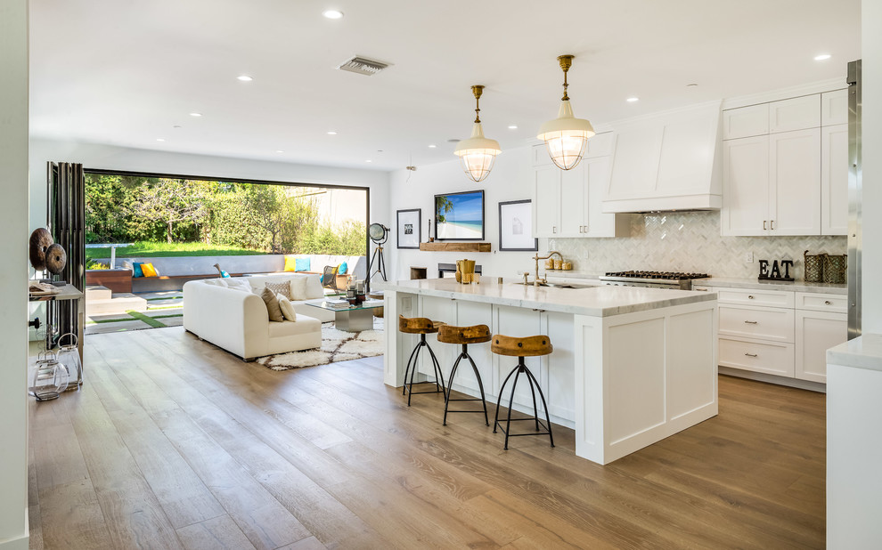 This is an example of a coastal l-shaped open plan kitchen in Los Angeles with shaker cabinets, white cabinets, white splashback, medium hardwood flooring, an island, brown floors and white worktops.