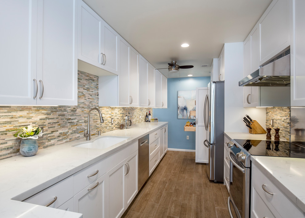 This is an example of a large classic galley kitchen pantry in Los Angeles with a submerged sink, shaker cabinets, white cabinets, engineered stone countertops, multi-coloured splashback, stone tiled splashback, stainless steel appliances, porcelain flooring, a breakfast bar and beige floors.