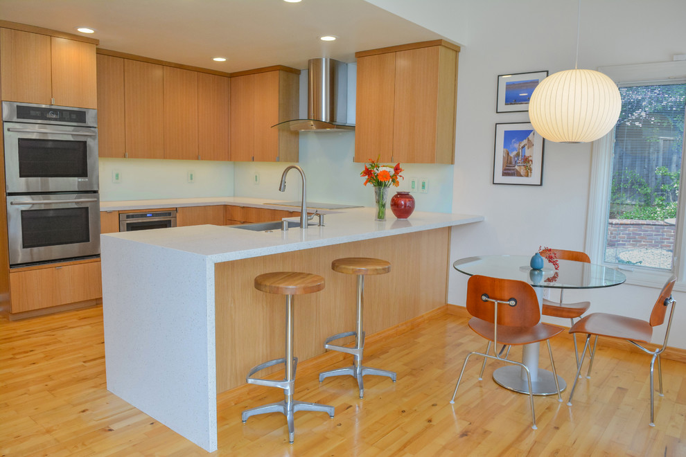 This is an example of a contemporary u-shaped kitchen/diner in San Francisco with flat-panel cabinets, light wood cabinets, quartz worktops, white splashback, glass sheet splashback, stainless steel appliances and light hardwood flooring.