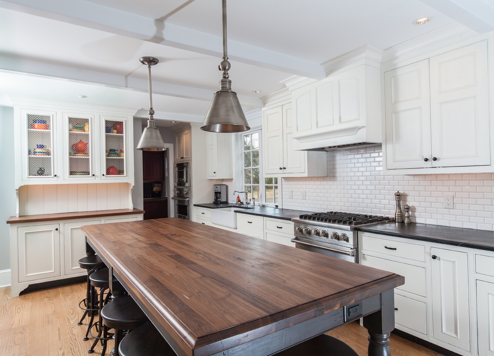 This is an example of a large traditional l-shaped kitchen/diner in Philadelphia with a single-bowl sink, white splashback, metro tiled splashback, stainless steel appliances, recessed-panel cabinets, white cabinets, light hardwood flooring, an island, brown floors, soapstone worktops and black worktops.