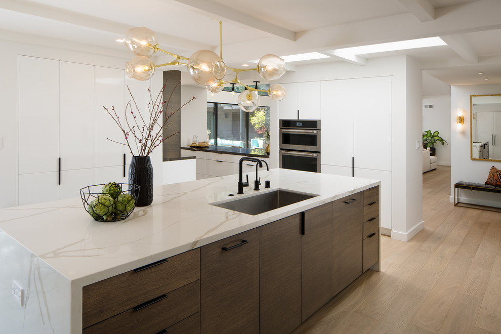 Bild på ett stort funkis vit vitt kök, med en undermonterad diskho, släta luckor, vita skåp, laminatbänkskiva, svart stänkskydd, stänkskydd i marmor, integrerade vitvaror, ljust trägolv, en köksö och brunt golv