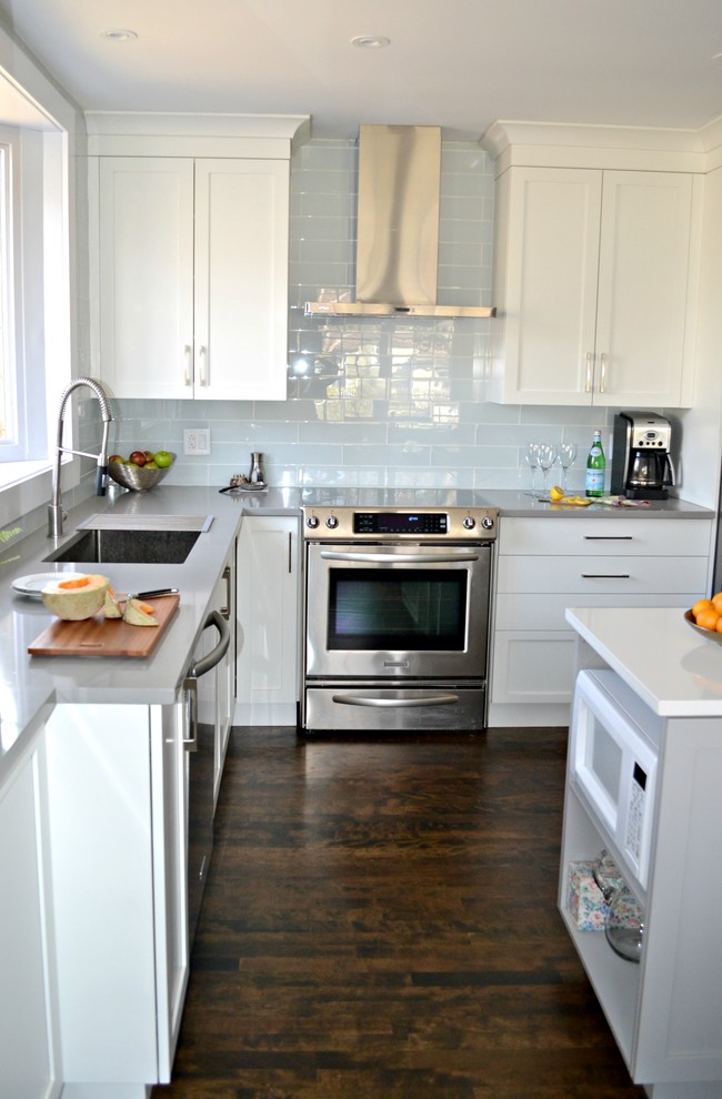 Foto på ett mellanstort funkis kök, med en undermonterad diskho, skåp i shakerstil, vita skåp, bänkskiva i kvarts, vitt stänkskydd, stänkskydd i glaskakel, rostfria vitvaror, mellanmörkt trägolv och en köksö