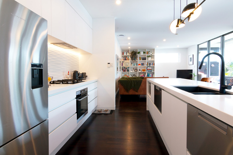 Contemporary galley kitchen in Auckland with a submerged sink, flat-panel cabinets, white cabinets, engineered stone countertops, white splashback, porcelain splashback, stainless steel appliances, dark hardwood flooring, an island, brown floors and white worktops.