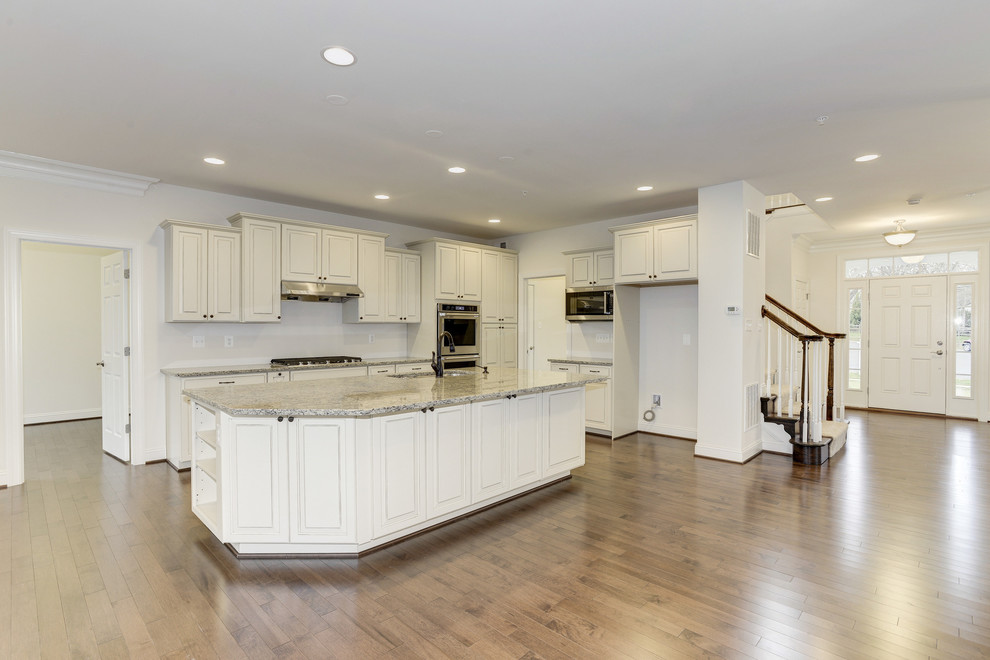 Traditional l-shaped open plan kitchen in DC Metro with a built-in sink, raised-panel cabinets, white cabinets, granite worktops, white splashback, stainless steel appliances, light hardwood flooring and an island.