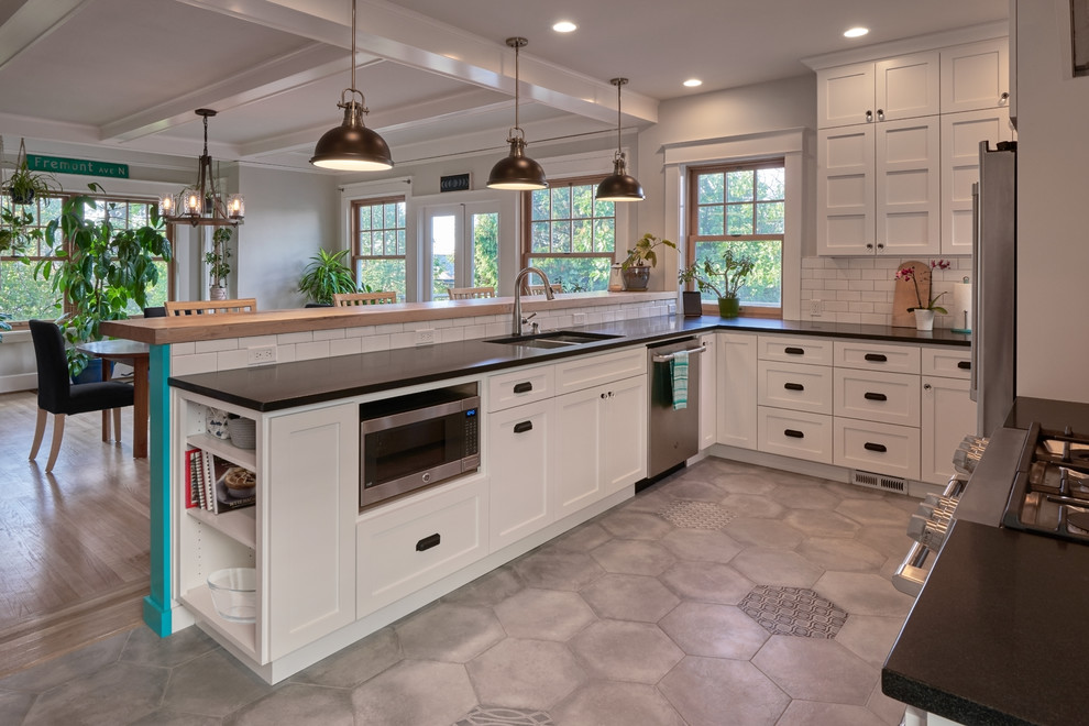 Photo of a medium sized farmhouse u-shaped kitchen/diner in Seattle with a double-bowl sink, shaker cabinets, white cabinets, granite worktops, white splashback, metro tiled splashback, stainless steel appliances, porcelain flooring, a breakfast bar, grey floors and black worktops.