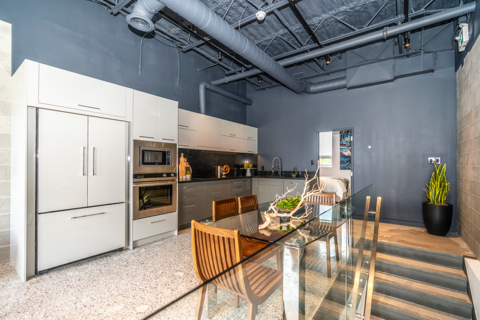 Photo of an industrial l-shaped open plan kitchen in Toronto with a submerged sink, flat-panel cabinets, white cabinets, black splashback, stone slab splashback, stainless steel appliances, no island, grey floors and black worktops.