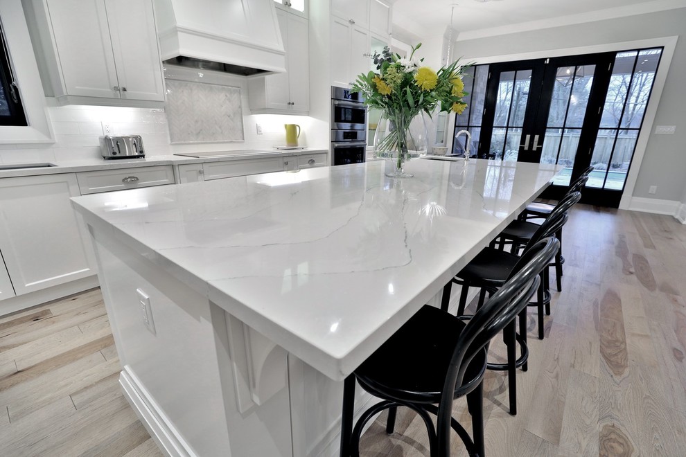 Mid-sized transitional l-shaped light wood floor eat-in kitchen photo with shaker cabinets, white cabinets, quartz countertops, white backsplash, glass tile backsplash, stainless steel appliances and an island