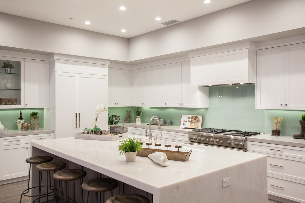 На фото: угловая кухня в стиле неоклассика (современная классика) с фасадами в стиле шейкер, белыми фасадами, зеленым фартуком, фартуком из стекла, островом и техникой под мебельный фасад