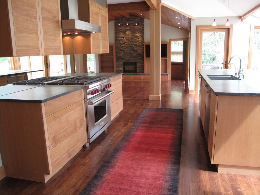 Cette image montre une cuisine ouverte parallèle design en bois clair avec un évier encastré, un placard à porte shaker, un plan de travail en granite, une crédence en feuille de verre, un électroménager en acier inoxydable et un sol en bois brun.