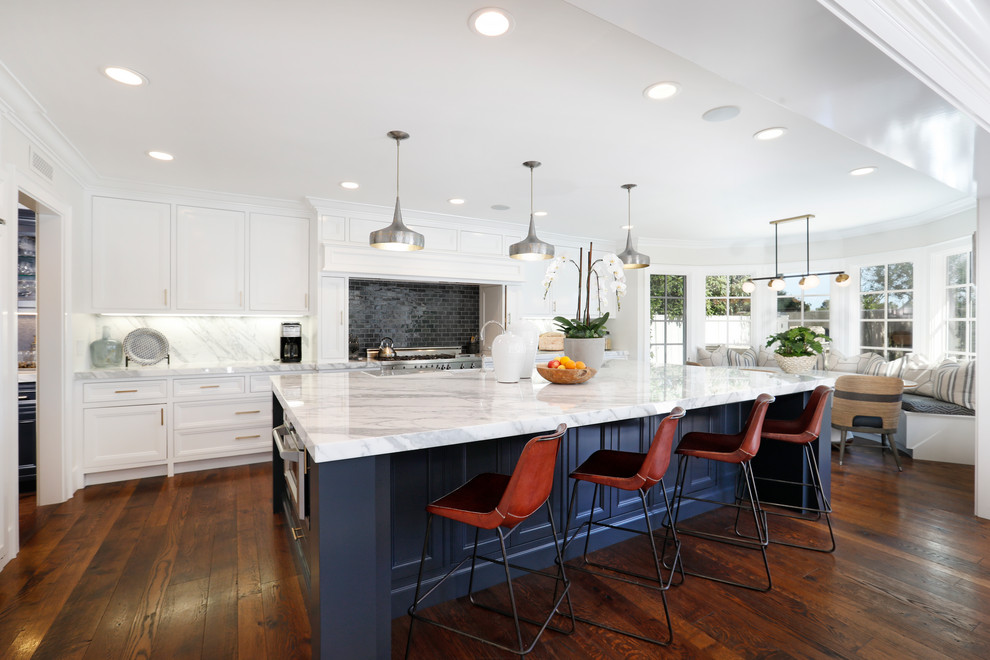 Example of a transitional galley medium tone wood floor and brown floor eat-in kitchen design in Orange County with a farmhouse sink, recessed-panel cabinets, white cabinets, gray backsplash, marble backsplash, stainless steel appliances, an island and gray countertops